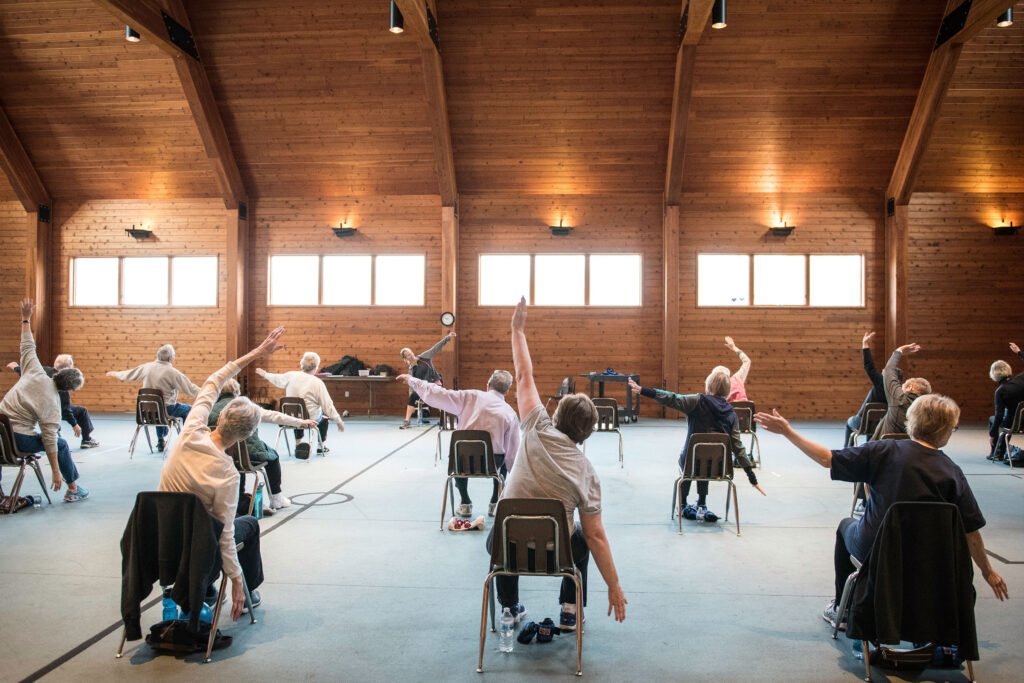 Rear view of gym filled with seniors exercising while seated in chairs led by instructor